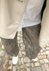Veste extérieure beige avec détails de boutons portée sur une chemise blanche froissée et un pantalon brun foncé avec des accents de coutures, assortis à des baskets blanches.