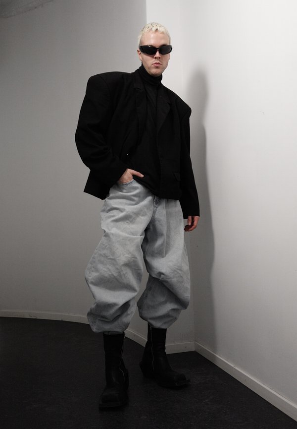 Persona con cabello corto y rubio, vistiendo un blazer negro oversize, suéter de cuello alto negro, pantalones claros y holgados, botas negras y gafas de sol oscuras, de pie en interiores.