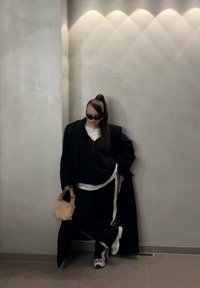 Abrigo negro de gran tamaño sobre una sudadera con capucha negra y pantalones con rayas blancas. Gafas de sol negras, bolso beige texturizado y zapatillas blancas. Fondo gris.