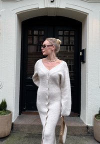 Γυναίκα με ξανθά μαλλιά, φορώντας λευκό μάλλινο φόρεμα με κουμπιά και γυαλιά ηλίου, στέκεται κρατώντας μια μπεζ τσάντα έξω από μια μαύρη πόρτα.