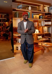 Homme portant un blazer en tweed marron clair, une chemise blanche, un jean large bleu marine, et des chaussures vertes, debout près d'une bibliothèque en bois avec un éclairage doux.