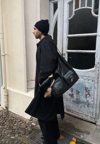 Čierny vlnený kabát, čierna pletená čiapka, kožená taška s kožušinovými detailmi a tmavé rukavice. V pozadí je textúrované, ošúchané dvere.