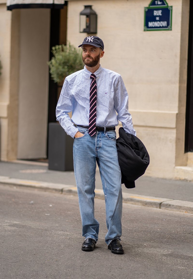 Homem com uma camisa de botão listrada em azul claro, uma gravata escura com listras rosas, jeans azul claro e sapatos pretos, segurando um casaco escuro.