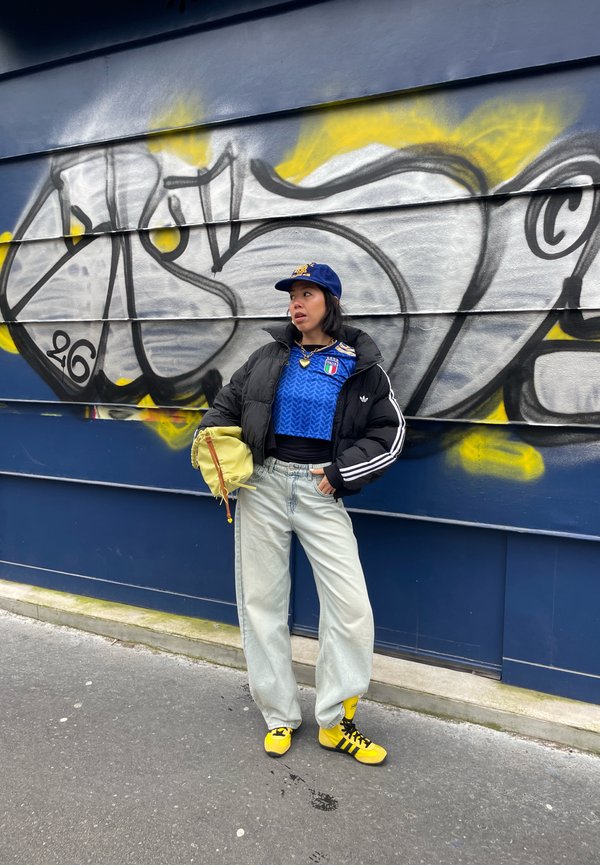 Junge Frau in blauem Sporttrikot, schwarzer Steppjacke, hellen Jeans, gelben Turnschuhen und Mütze steht vor einer mit Graffiti bedeckten Wand.