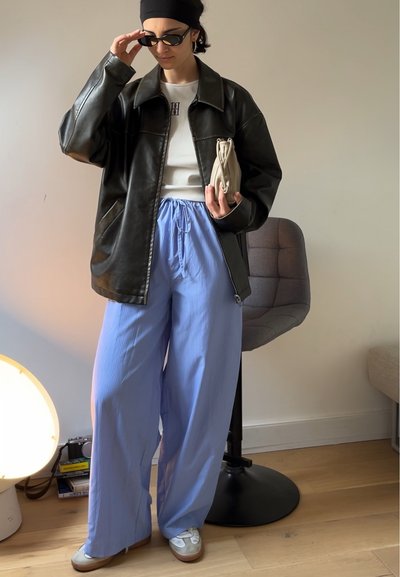 Chaqueta de cuero negra de gran tamaño, camisa blanca ajustada, pantalones anchos de rayas finas en azul claro y zapatos grises. Incluye un bolso de mano beige y gafas de sol.