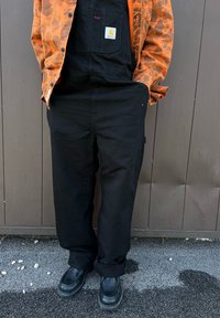 Μαύρες φόρμες φτιαγμένες από ανθεκτικό ύφασμα, με μια μπροστινή τσέπη που έχει ετικέτα μάρκας. Συνδυασμένες με ένα πορτοκαλί πανωφόρι με σχέδια και μαύρα παπούτσια.