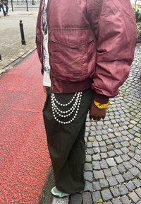 Osoba w bordowej kurtce, oliwkowych spodniach, z warstwami pereł na łańcuchach, żółtym zegarkiem, stojąca na kostce brukowej i czerwonym chodniku.