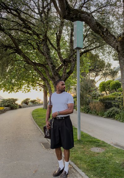 Hombre con camiseta blanca y pantalones cortos negros está de pie en un camino curvado bajo grandes árboles frondosos, sosteniendo una bolsa y mirando hacia un lado cerca del cartel de Montreux.