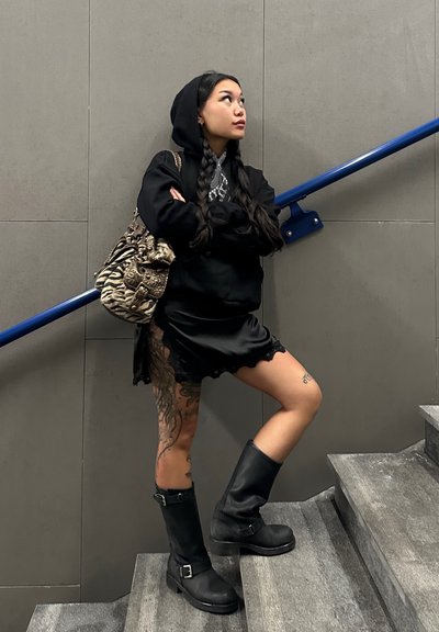 Modelo vistiendo un hoodie negro y un vestido negro de satén con dobladillo de encaje, botas hasta la rodilla negras y un bolso de hombro estampado, de pie en unas escaleras.