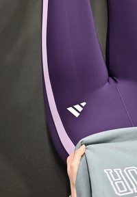Vêtement de sport violet avec des bandes latérales rose clair et un détail logo blanc. Le tissu semble extensible et lisse sur un fond sombre.
