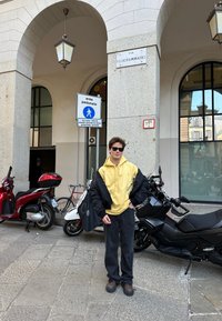 Oseba v rumenem kapucinem in črni jakni stoji blizu skuterjev, v ozadju pa je znak, ki označuje pešcono cono.