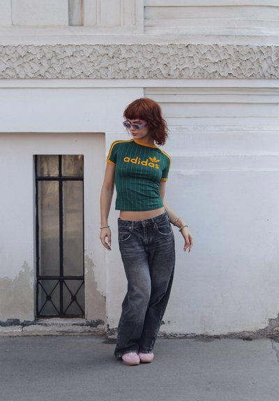 Top corto a rayas verdes y amarillas con el logo de "adidas", combinado con jeans grises holgados y calzado rosa, todo contra un fondo blanco.