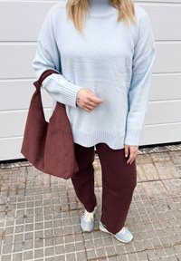Personne portant un pull bleu clair et un pantalon marron tenant un grand sac à main en daim marron, debout sur un trottoir carrelé devant une porte blanche.
