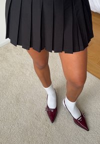 Schwarzer plissierter Rock mit kurzem Saum, kombiniert mit weißen Socken und kastanienbraunen lackierten spitzen Ballerinas mit Schnallenakzenten.