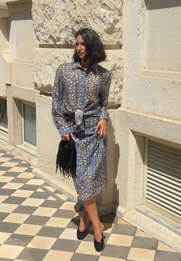 Femme aux cheveux courts et foncés portant un chemisier floral semi-transparent et une jupe assortie, tenant un sac noir à franges, debout sur un trottoir à carreaux.