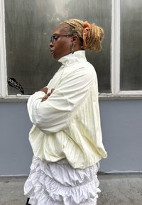 Casaco plissado amarelo claro com gola alta e mangas oversize; saia branca com folhos em camadas; brincos grandes e elástico de cabelo como accentos.