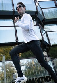 T-shirt de corrida de mangas longas brancas com leggings ajustados pretos e sapatilhas atléticas brancas com riscas pretas; o fundo inclui arquitetura em vidro.