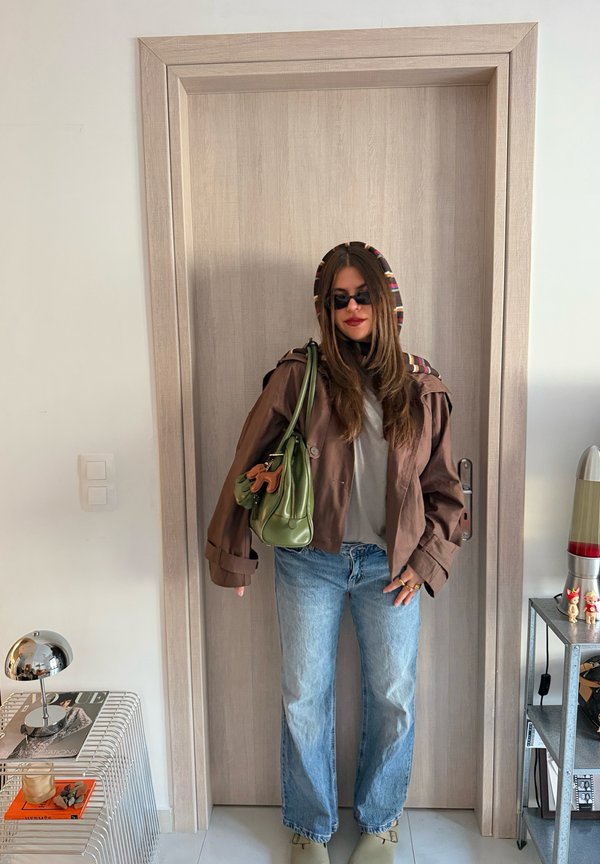 Une femme pose devant une porte en bois, portant des lunettes de soleil, une veste marron et un jean bleu, avec un sac vert en bandoulière.