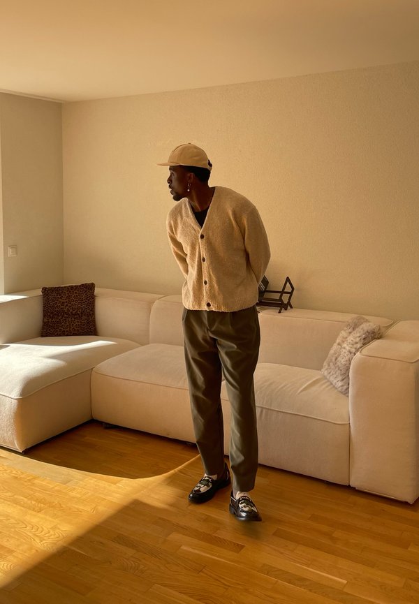 Uomo con cardigan beige, cappello e pantaloni scuri in piedi vicino a un divano crema con cuscini in un soggiorno illuminato dal sole con pavimento in legno.