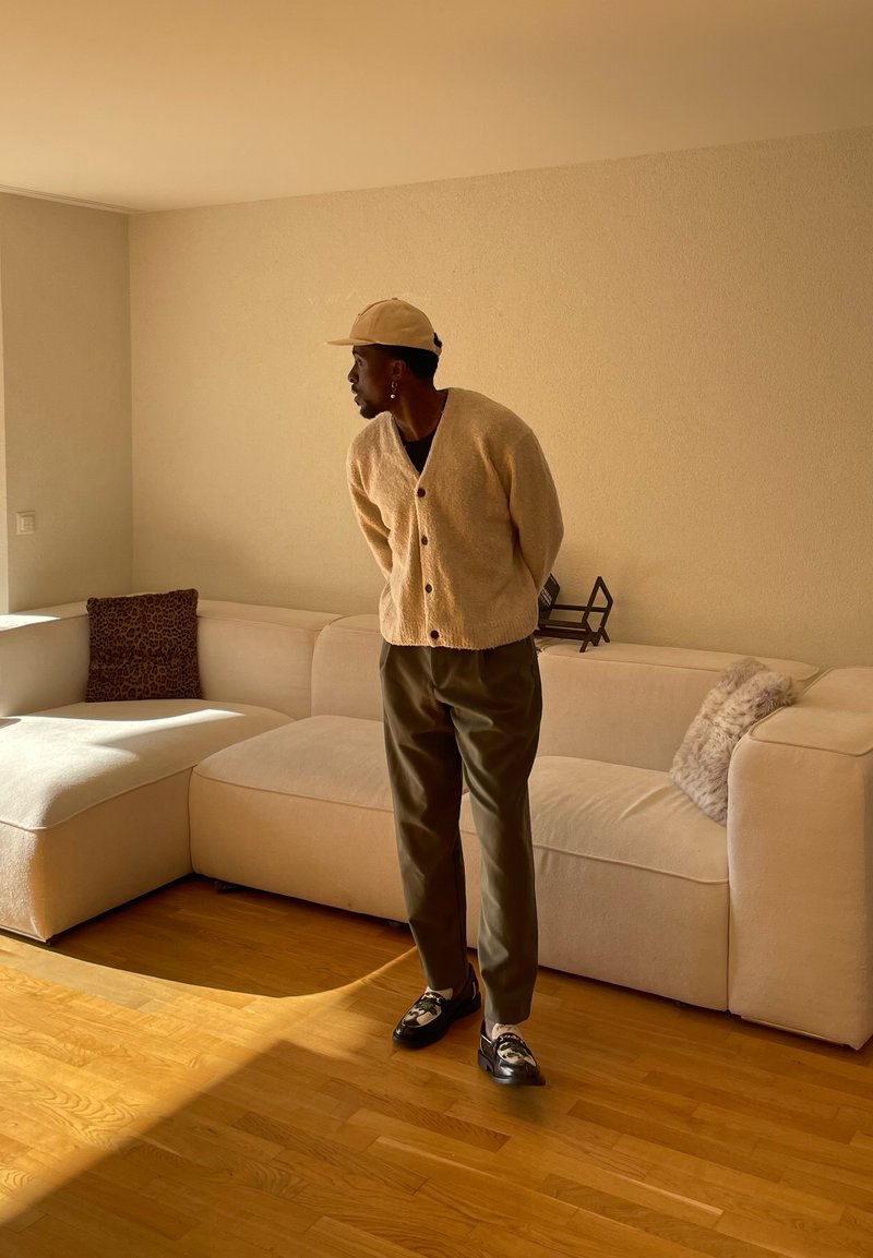 Homme en cardigan beige, casquette et pantalon sombre, debout près d'un canapé crème avec coussins dans un salon baigné de lumière avec un sol en bois.