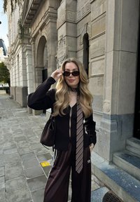 Tumša garroku augšdaļa kombinēta ar platu kāju bordo sporta biksēm ar baltām svītrām. Aksesuāri ietver rakstainu kaklasaiti un saulesbrilles.