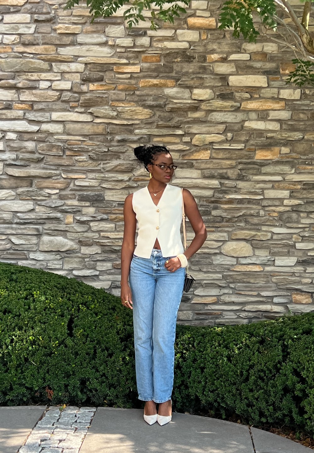 Λευκό αμάνικο τοπ με V λαιμόκοψη και κουμπιά, σε συνδυασμό με μπλε τζιν ίσιας γραμμής και λευκά μυτερά τακούνια, μπροστά από έναν πέτρινο τοίχο.