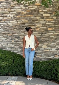 Λευκό αμάνικο τοπ με V λαιμόκοψη και κουμπιά, σε συνδυασμό με μπλε τζιν ίσιας γραμμής και λευκά μυτερά τακούνια, μπροστά από έναν πέτρινο τοίχο.