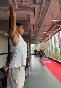 Homme étirant les triceps avec une bande de résistance dans une salle de sport, debout près d'une fenêtre avec des appareils d'exercice et une piste rouge à côté de lui.
