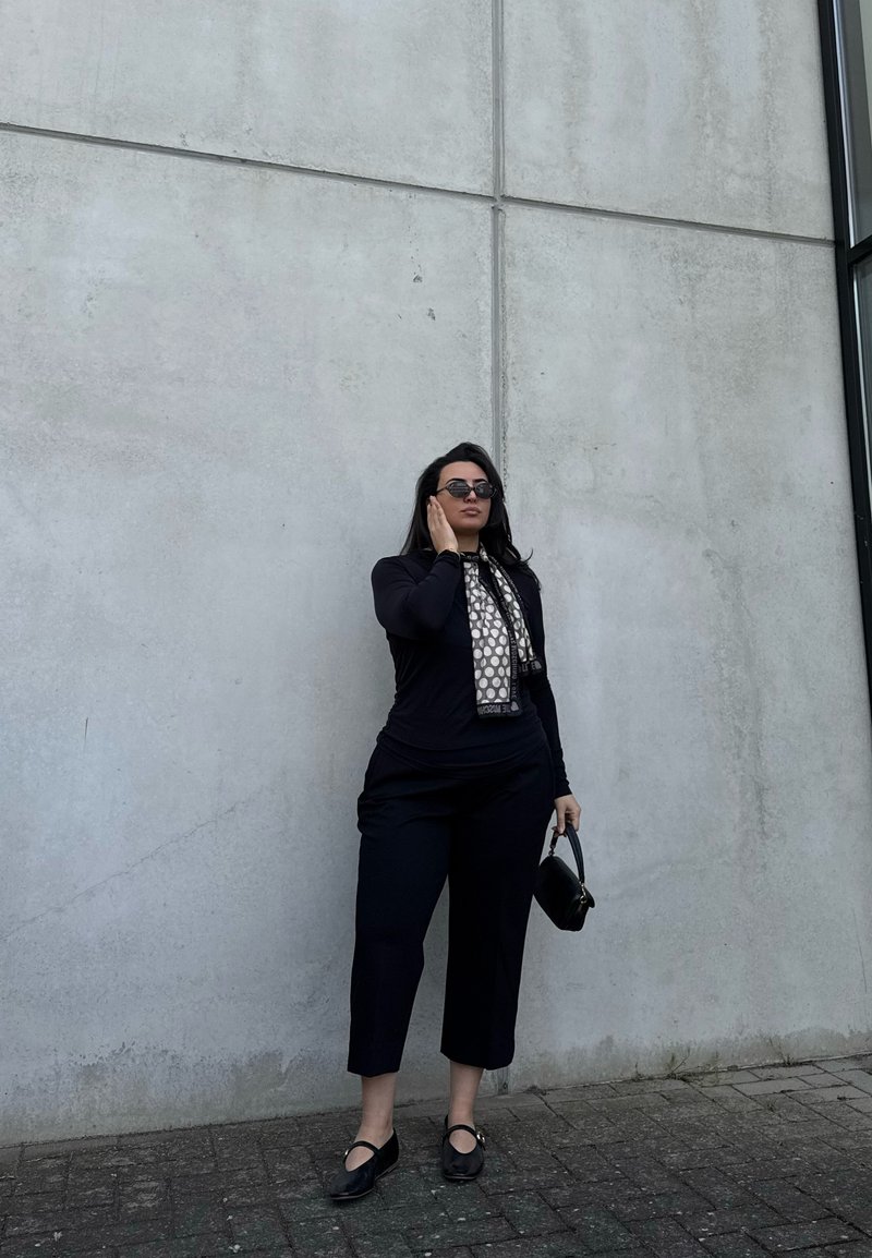 Woman wearing black outfit and sunglasses stands against gray concrete wall, holding small black handbag and touching ear.