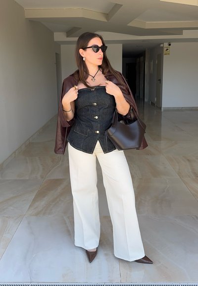 Mujer con gafas de sol, top oscuro sin tirantes, pantalones blancos y chaqueta marrón sobre los hombros, sosteniendo un bolso marrón en interiores.