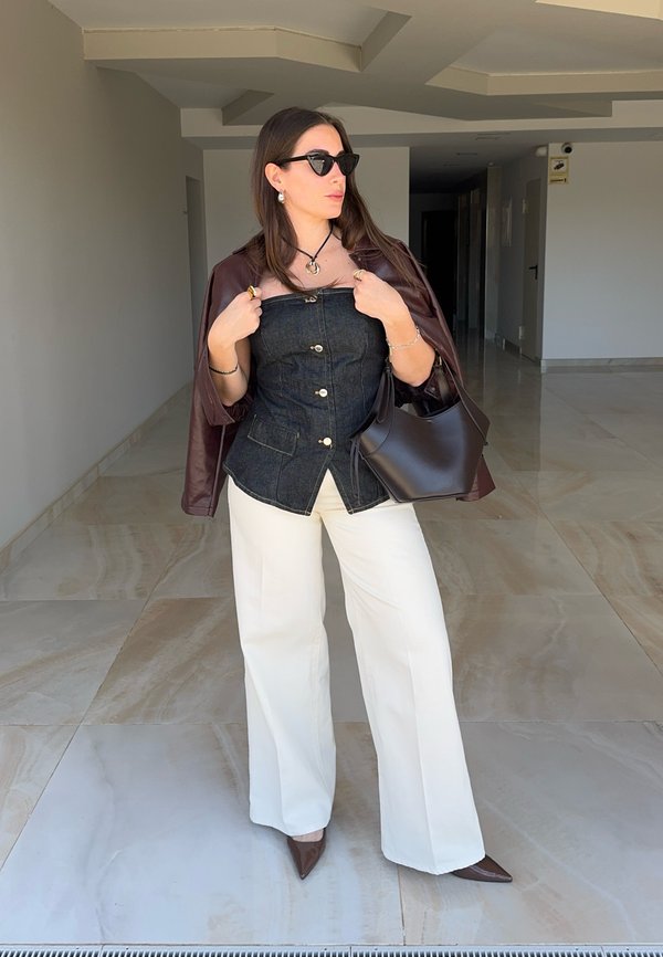 Donna con occhiali da sole, top scuro senza spalline, pantaloni bianchi e giacca marrone sulle spalle, che tiene una borsa marrone al chiuso.
