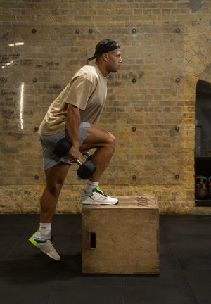 Black dumbbells held in one hand, worn beige t-shirt, gray shorts, and white sneakers with green accents; stepping onto a wooden box.