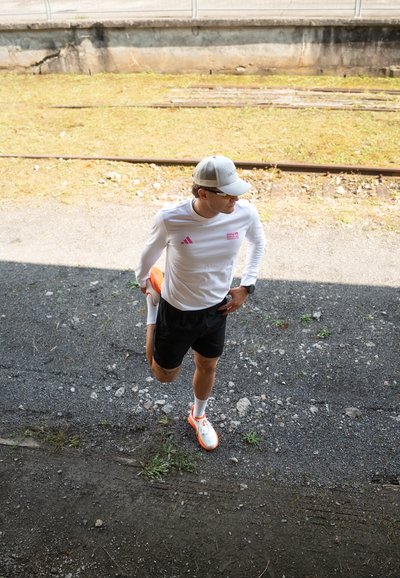 Atleta estirándose, vistiendo una camiseta de manga larga blanca con detalles en rosa, pantalones cortos negros y zapatillas de correr en naranja y blanco. Gorra gris y gafas de sol.