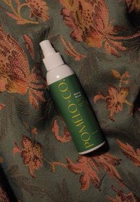 Grøn sprayflaske mærket "Pomelo + Co. Matcha Moves" på en struktureret, blomsterprintet stofbaggrund.