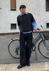 Camicia nera a maniche corte sopra una camicia celeste chiaro con colletto, abbinata a jeans di jeans scuri e larghi e scarpe nere, appoggiata a una bicicletta.