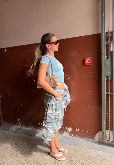 Joven mujer con cabello largo recogido en una coleta, llevando gafas de sol, top corto azul, pantalones cargo de camuflaje, chanclas y llevando un bolso beige de hombro, está de pie en el interior.