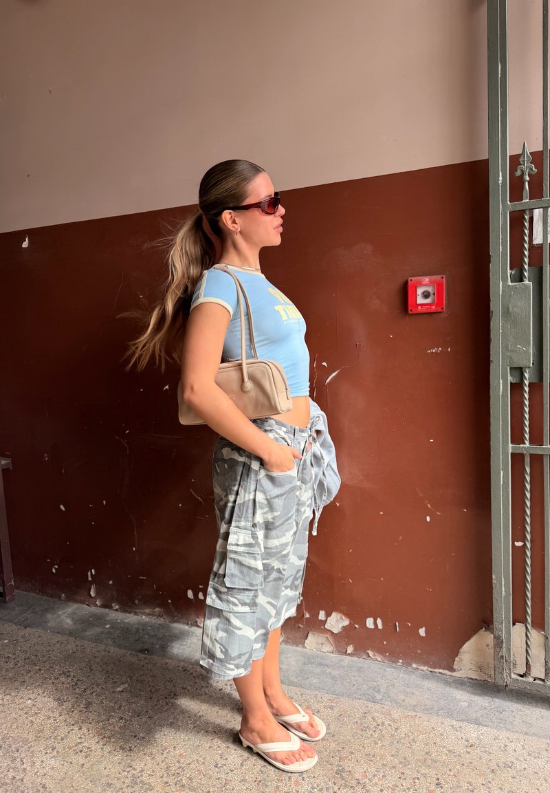 Jeune femme aux longs cheveux attachés en queue de cheval, portant des lunettes de soleil, un crop top bleu, un pantalon cargo camouflage, des tongs et un sac à bandoulière beige, se tient à l'intérieur.