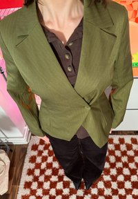 Personne portant un blazer à fines rayures vert olive sur une chemise marron foncé boutonnée, un pantalon noir et des chaussures noires pointues, debout sur un tapis à carreaux rouge et blanc.
