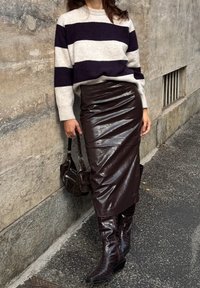 Persona che indossa un maglione a righe bianche e nere, una gonna di pelle lucida marrone scuro, stivali abbinati, mentre tiene una borsa nera, in piedi accanto a un muro di pietra.