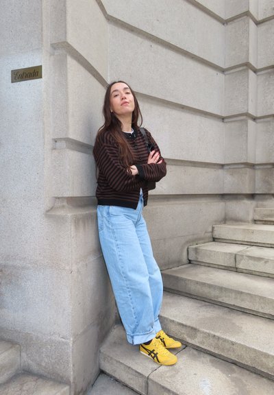 Joven con pelo largo y castaño, que lleva un suéter a rayas, vaqueros azul claro y zapatillas amarillas, de pie junto a unas escaleras de piedra cerca de un cartel de entrada.