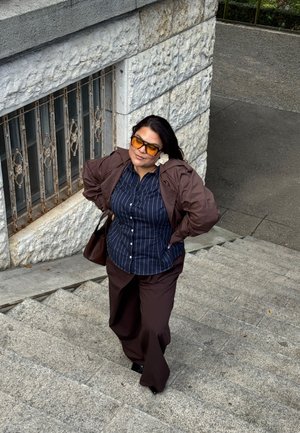 Veste marron sur une chemise rayée bleue, associée à un pantalon marron. Les lunettes de soleil ont des verres teintés jaunes. Escaliers en pierre et un mur texturé en arrière-plan.