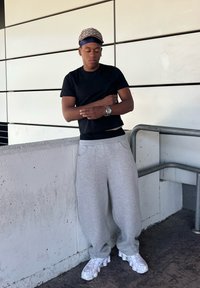 Black short-sleeved shirt, grey sweatpants with pockets, white trainers, and a patterned cap. He stands against a wall and railing.
