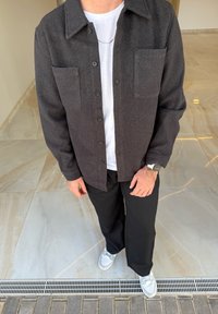 Camisa cinza de botão em tecido texturizado com dois bolsos no peito, sobre uma t-shirt branca. Calças pretas e ténis cinzentos completam o look.