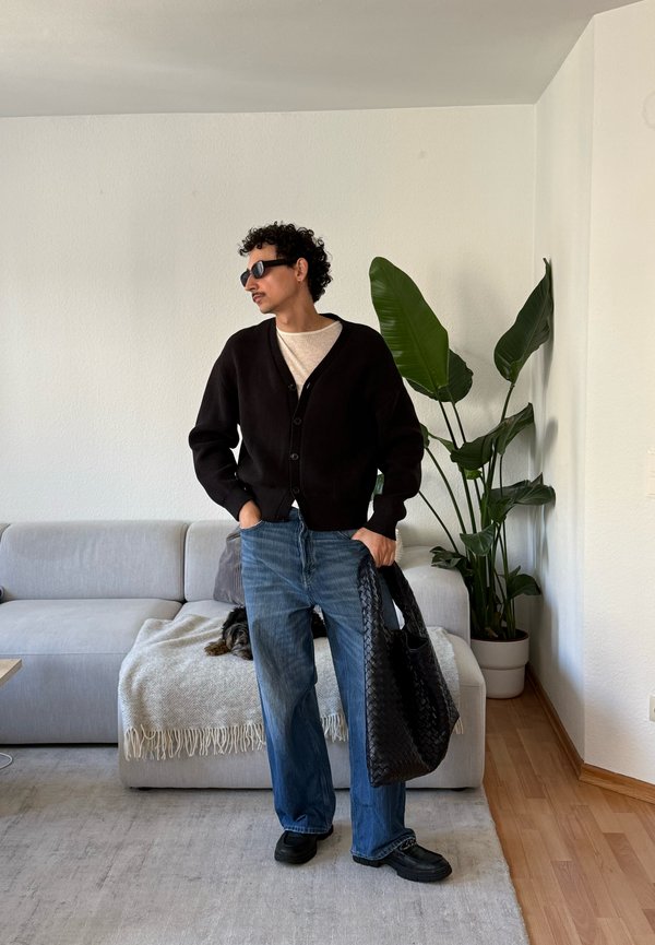 Mand iført sort cardigan, beige skjorte, blå jeans og solbriller står indendørs med en sort flettet taske nær en sofa og en stor potteplante.