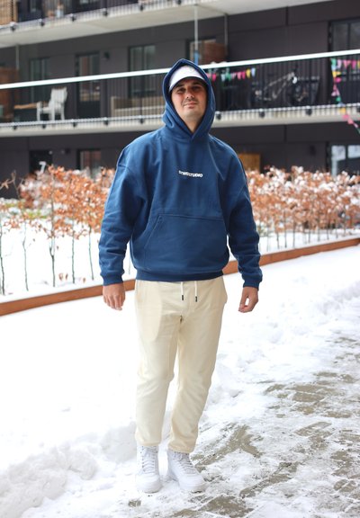 Hombre con sudadera azul, gorra blanca, pantalones beige y botas blancas de pie en un camino nevado frente a un edificio de apartamentos moderno.