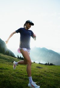 Kvinne som løper i en blå skjorte og lyse lilla shorts, iført solhatt og hvite sko med røde detaljer, løper på gressdekket terrain.
