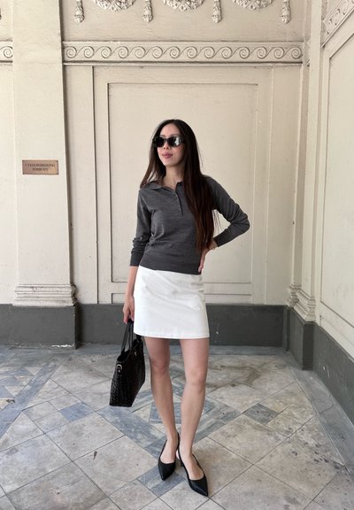 Mujer con el pelo largo que lleva gafas de sol, camiseta gris de manga larga, falda blanca y tacones negros, sosteniendo un bolso negro en el interior sobre un suelo de baldosas.