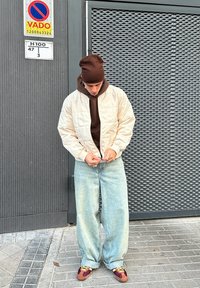 Beige gesteppte Bomberjacke über einem braunen Kapuzenpullover, mit hellblauen Jeans mit weitem Bein und braunen sowie burgunderfarbenen Sneakers. Strukturierter Hintergrund.