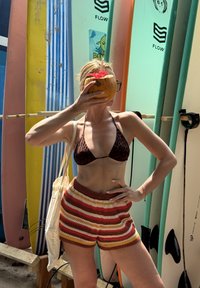 Vrouw in een bikinibovenstuk met luipaardprint en gestreepte korte broek, die een kokosnootdrank vasthoudt en voor kleurrijke surfplanken staat.