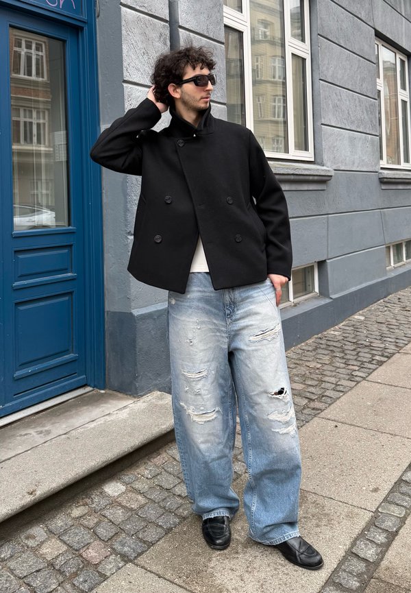 Bărbat purtând o jachetă neagră cu două rânduri de nasturi, blugi largi cu rupturi, pantofi negri și ochelari de soare, stă pe trotuarul de piatră cubică lângă o ușă albastră și ferestre.